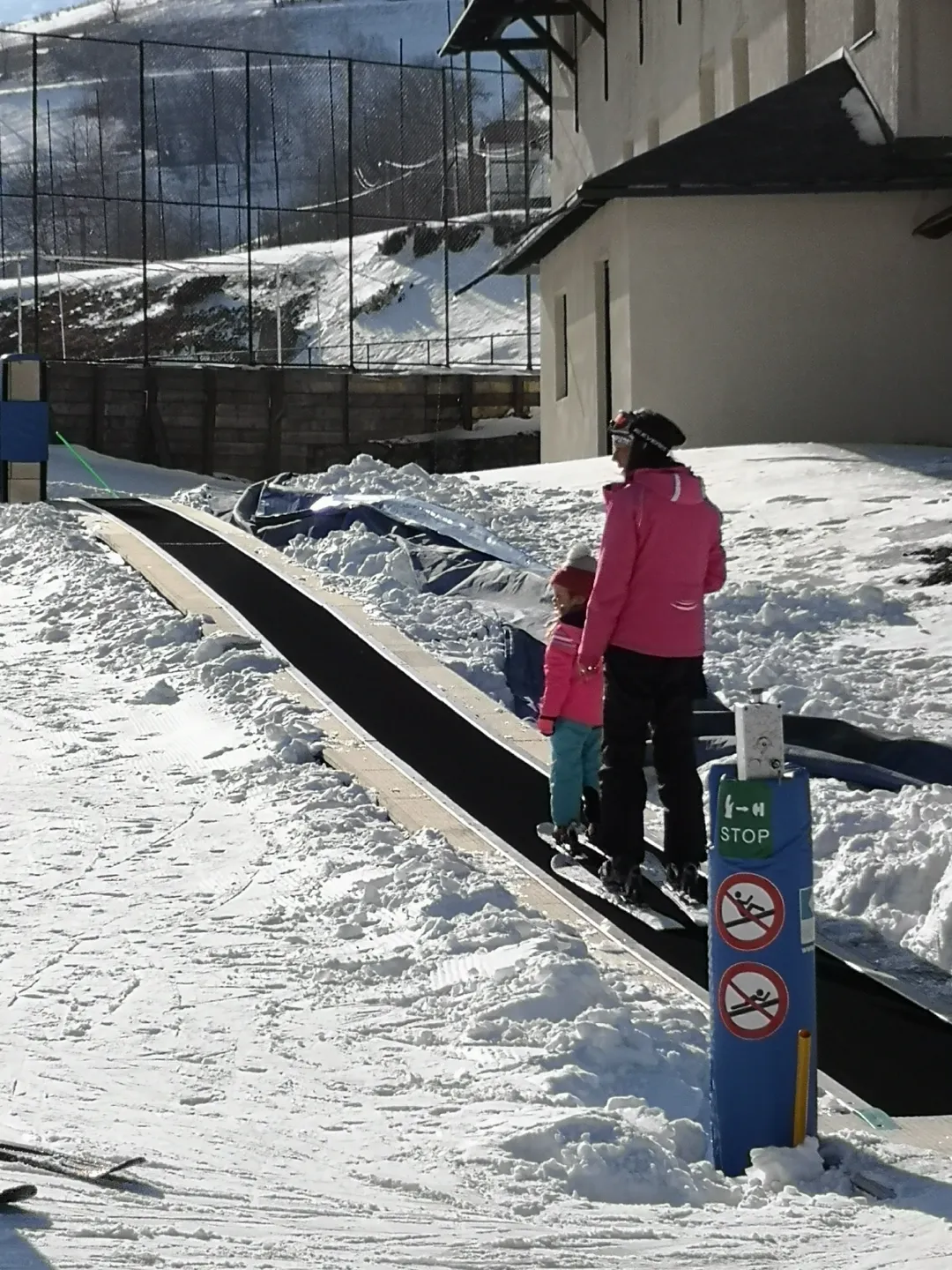 A child and an adult on skis standing on the magic carpet as it carries them up the slope